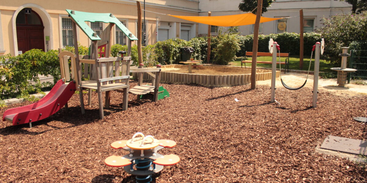 Kinderspielplatz mit Klettergerüst, Rutsche, Sandkasten mit Überdachung, Springreiter, Schaukel und Bänken, auf Mulch in einem von Gebäuden umgebenen Innenhof gelegen.