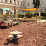 Kinderspielplatz mit Klettergerüst, Rutsche, Sandkasten mit Überdachung, Springreiter, Schaukel und Bänken, auf Mulch in einem von Gebäuden umgebenen Innenhof gelegen.