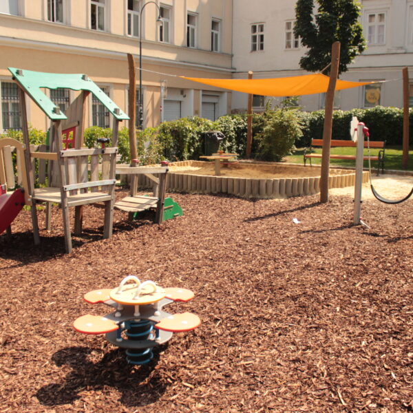 Kinderspielplatz mit Klettergerüst, Rutsche, Sandkasten mit Überdachung, Springreiter, Schaukel und Bänken, auf Mulch in einem von Gebäuden umgebenen Innenhof gelegen.