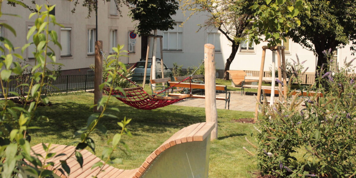 Ein kleiner Stadtpark mit einer modernen Holzbank, einer roten Hängematte, grünem Gras, Bäumen und nahe gelegenen Wohnhäusern.