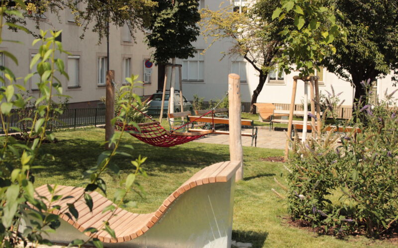 Ein kleiner Stadtpark mit einer modernen Holzbank, einer roten Hängematte, grünem Gras, Bäumen und nahe gelegenen Wohnhäusern.