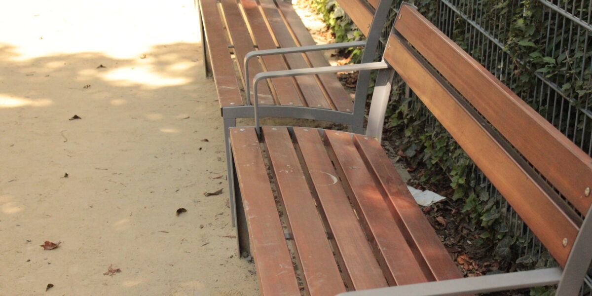 Drei leere Holzbänke mit Metallarmlehnen stehen auf einem unbefestigten Weg neben einem mit grünem Efeu bewachsenen Metallzaun.