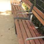 Drei leere Holzbänke mit Metallarmlehnen stehen auf einem unbefestigten Weg neben einem mit grünem Efeu bewachsenen Metallzaun.