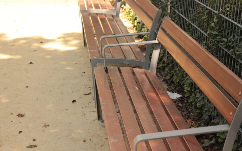 Drei leere Holzbänke mit Metallarmlehnen stehen auf einem unbefestigten Weg neben einem mit grünem Efeu bewachsenen Metallzaun.