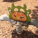 Eine gefederte Spielplatzwippe mit grünem und orangefarbenem Insektenmotiv auf einer mulchbedeckten Oberfläche.