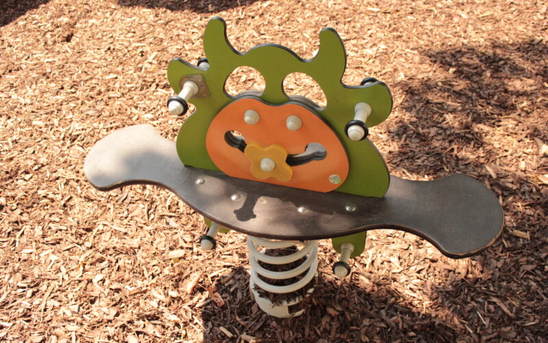 Eine gefederte Spielplatzwippe mit grünem und orangefarbenem Insektenmotiv auf einer mulchbedeckten Oberfläche.