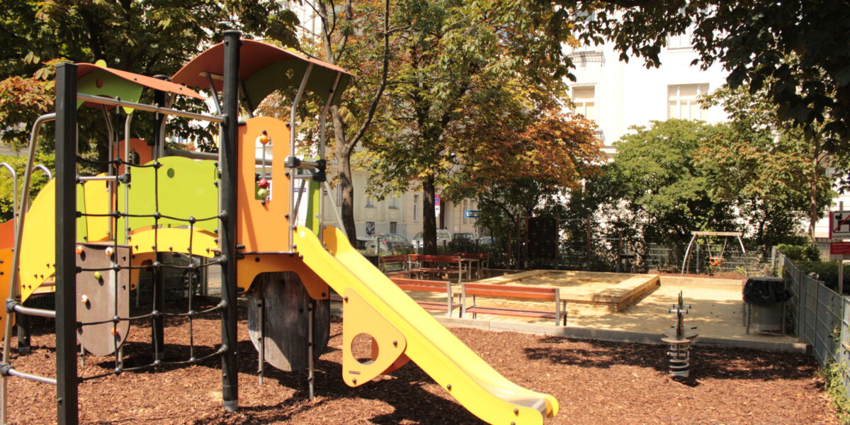 Ein Spielplatz im Freien mit einer gelben Rutsche, einem Klettergerüst, Bänken, einem Sandkasten und einer Schaukel, umgeben von Bäumen und Holzspänen unter den Füßen.