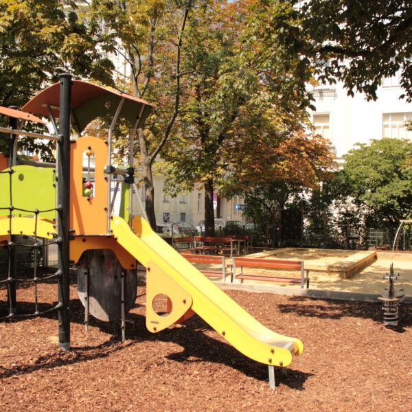 Ein Spielplatz im Freien mit einer gelben Rutsche, einem Klettergerüst, Bänken, einem Sandkasten und einer Schaukel, umgeben von Bäumen und Holzspänen unter den Füßen.