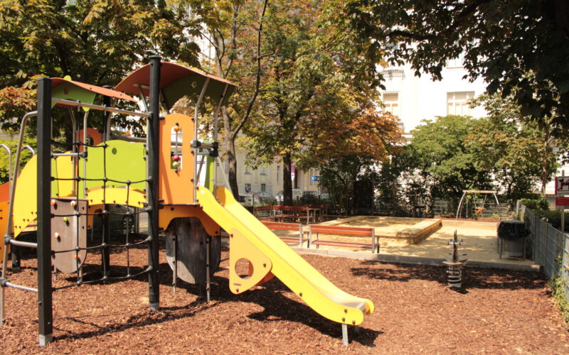 Ein Spielplatz im Freien mit einer gelben Rutsche, einem Klettergerüst, Bänken, einem Sandkasten und einer Schaukel, umgeben von Bäumen und Holzspänen unter den Füßen.