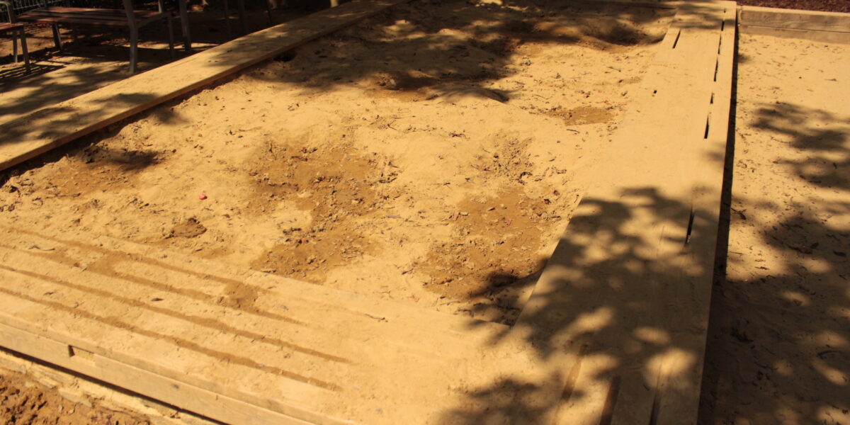 Ein großer rechteckiger Sandkasten mit hölzernen Umrandungen, teilweise im Schatten von Bäumen, mit unebenem Sand und Fußabdrücken.