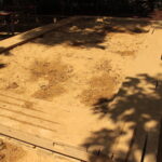 Ein großer rechteckiger Sandkasten mit hölzernen Umrandungen, teilweise im Schatten von Bäumen, mit unebenem Sand und Fußabdrücken.