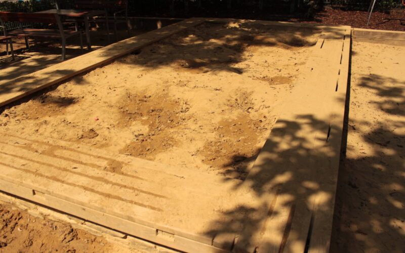 Ein großer rechteckiger Sandkasten mit hölzernen Umrandungen, teilweise im Schatten von Bäumen, mit unebenem Sand und Fußabdrücken.
