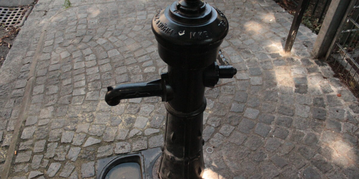 Eine öffentliche Wasserpumpe aus schwarzem Gusseisen steht auf einem Kopfsteinpflaster, neben einem Metallzaun und einem Abflussgitter.