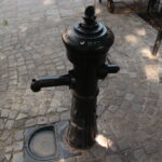Eine öffentliche Wasserpumpe aus schwarzem Gusseisen steht auf einem Kopfsteinpflaster, neben einem Metallzaun und einem Abflussgitter.
