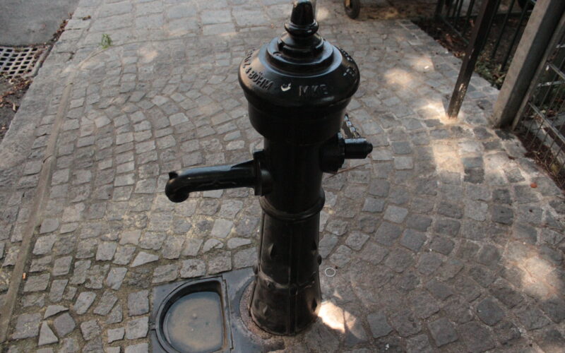 Eine öffentliche Wasserpumpe aus schwarzem Gusseisen steht auf einem Kopfsteinpflaster, neben einem Metallzaun und einem Abflussgitter.