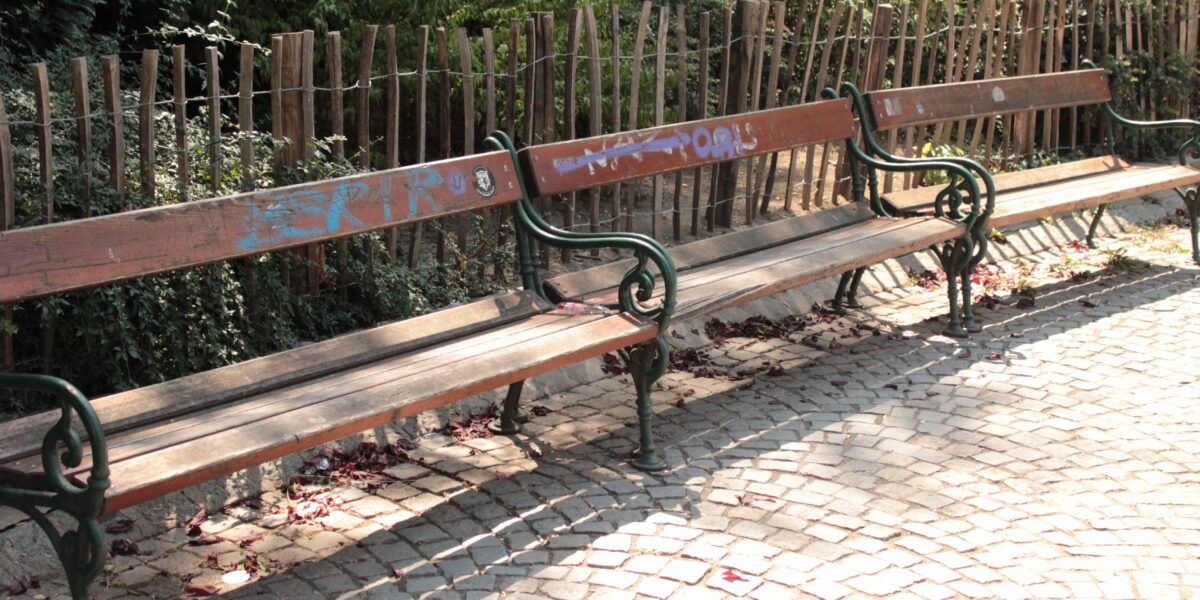 Drei leere Holzbänke mit grünen Metallrahmen stehen entlang eines gepflasterten Weges, mit einem Holzzaun und Grünpflanzen im Hintergrund. Auf den Bänken sind einige Graffiti zu sehen.