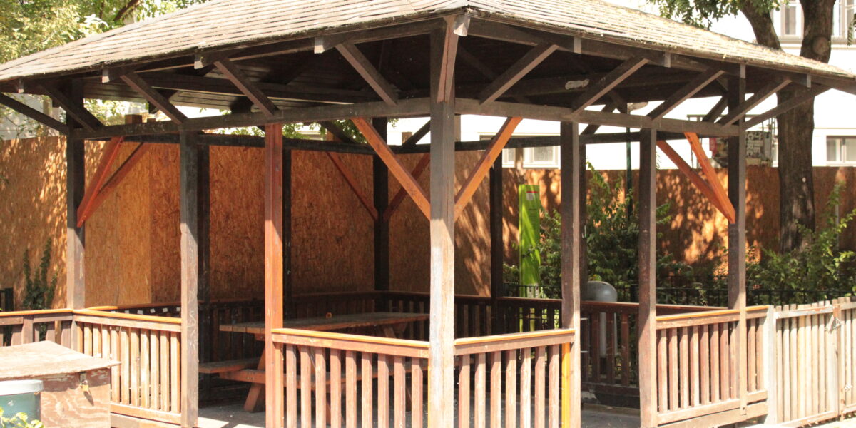 Ein hölzerner Pavillon mit Spitzdach und offenen Seiten steht auf einer gepflasterten Fläche, umgeben von Bäumen und einem Holzzaun.