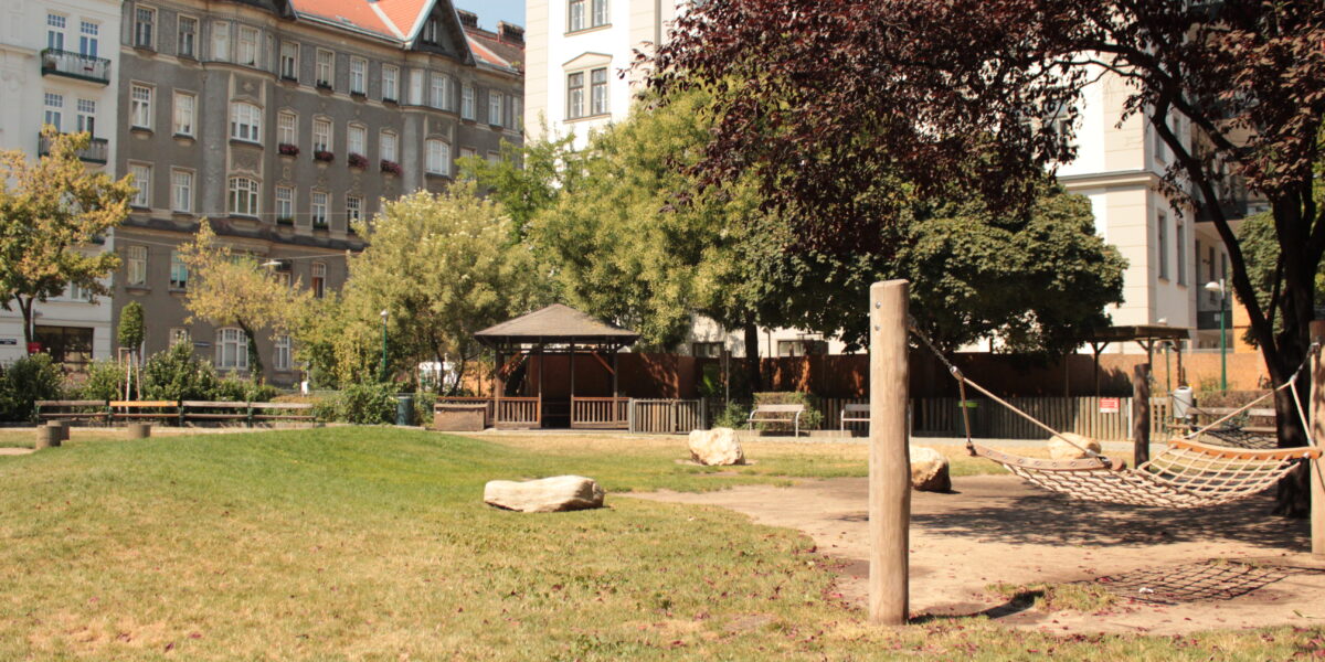 Ein Stadtpark mit Hängematte, grünem Rasen, großen Felsen, Bäumen und einem Pavillon, umgeben von mehrstöckigen Wohngebäuden.