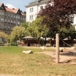 Ein Stadtpark mit Hängematte, grünem Rasen, großen Felsen, Bäumen und einem Pavillon, umgeben von mehrstöckigen Wohngebäuden.