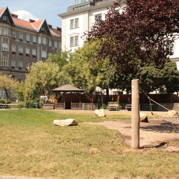 Ein Stadtpark mit Hängematte, grünem Rasen, großen Felsen, Bäumen und einem Pavillon, umgeben von mehrstöckigen Wohngebäuden.