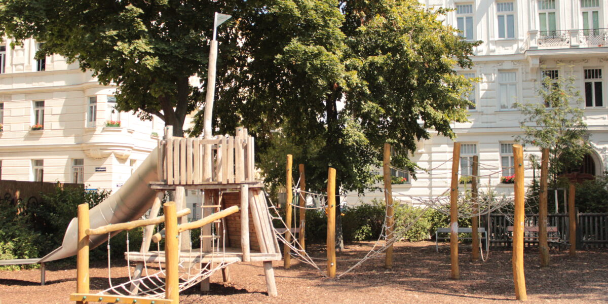 Ein Spielplatz aus Holz mit Seilbrücken und einer Rutsche befindet sich in einem Park, umgeben von Bäumen und angrenzend an weiße Wohnhäuser.