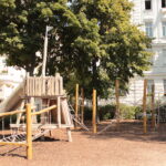 Ein Spielplatz aus Holz mit Seilbrücken und einer Rutsche befindet sich in einem Park, umgeben von Bäumen und angrenzend an weiße Wohnhäuser.