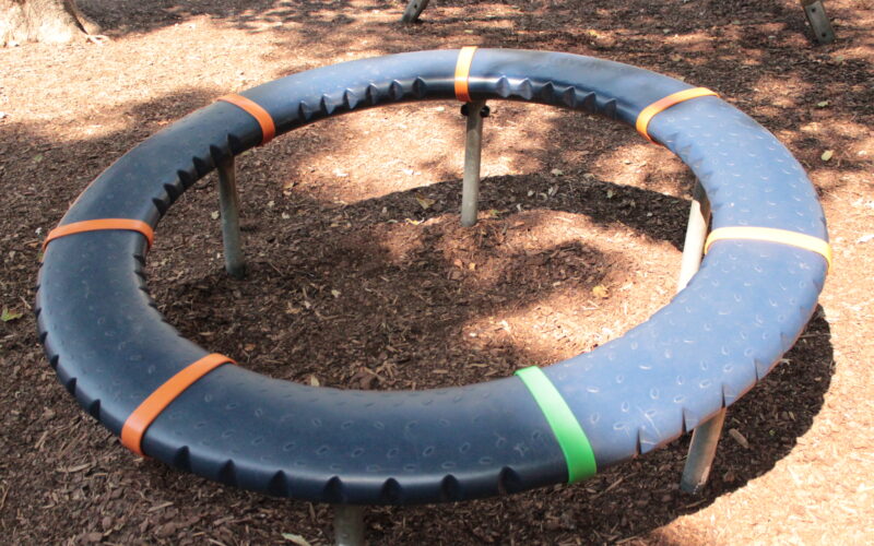 Ein großes kreisförmiges blaues Spielgerät mit orangefarbenen und grünen Teilen, das von Metallfüßen getragen wird, steht auf einem mit Rinde bedeckten Boden im gedämpften Sonnenlicht.