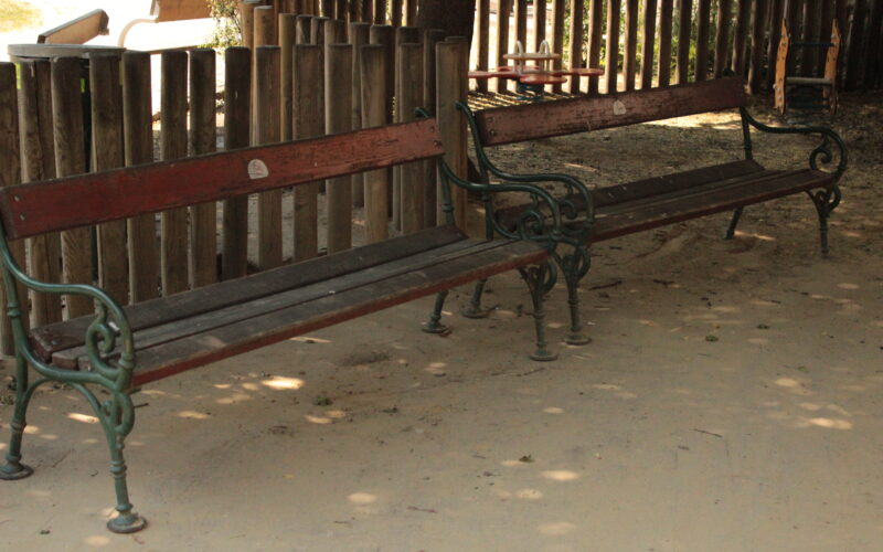 Zwei leere Holzbänke mit Metallarmlehnen stehen auf einem unbefestigten Boden in der Nähe eines Holzzauns in einem Außenbereich.