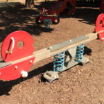Rote und graue Wippe mit Federstützen auf einem Spielplatz mit Mulchbodenbelag, bei Tageslicht fotografiert.