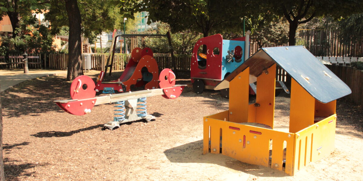 Ein Spielplatz mit einer Federwippe, einem Spielhaus und Klettergerüsten, umgeben von Bäumen und einem Holzzaun an einem sonnigen Tag.