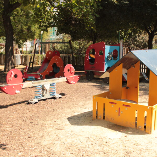 Ein Spielplatz mit einer Federwippe, einem Spielhaus und Klettergerüsten, umgeben von Bäumen und einem Holzzaun an einem sonnigen Tag.