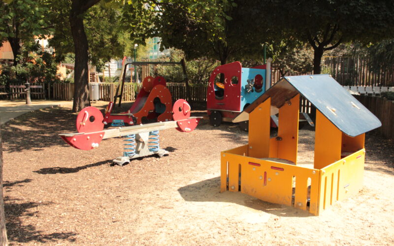 Ein Spielplatz mit einer Federwippe, einem Spielhaus und Klettergerüsten, umgeben von Bäumen und einem Holzzaun an einem sonnigen Tag.