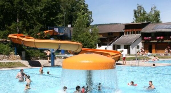 Menschen schwimmen und spielen in einem Außenpool mit einem pilzförmigen Springbrunnen und einer gelben Wasserrutsche im Hintergrund.
