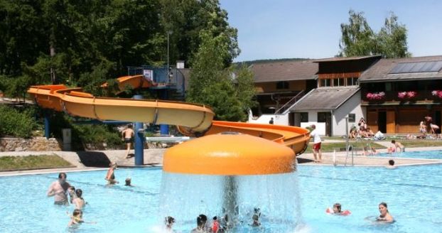 Menschen schwimmen und spielen in einem Außenpool mit einem pilzförmigen Springbrunnen und einer gelben Wasserrutsche im Hintergrund.
