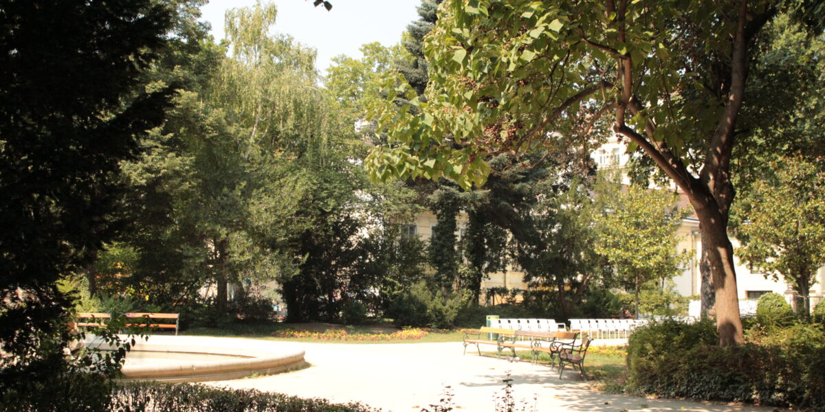 Sonnenbeschienener Park mit leeren Bänken, belaubten Bäumen und einer runden gepflasterten Fläche in der Mitte; dichtes Grün umgibt die Freifläche.