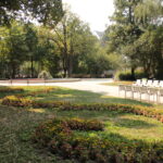 Eine angelegte Parkanlage mit geschwungenen Blumenbeeten, in Reihen angeordneten leeren weißen Stühlen und Bäumen, die an einem sonnigen Tag Schatten spenden.