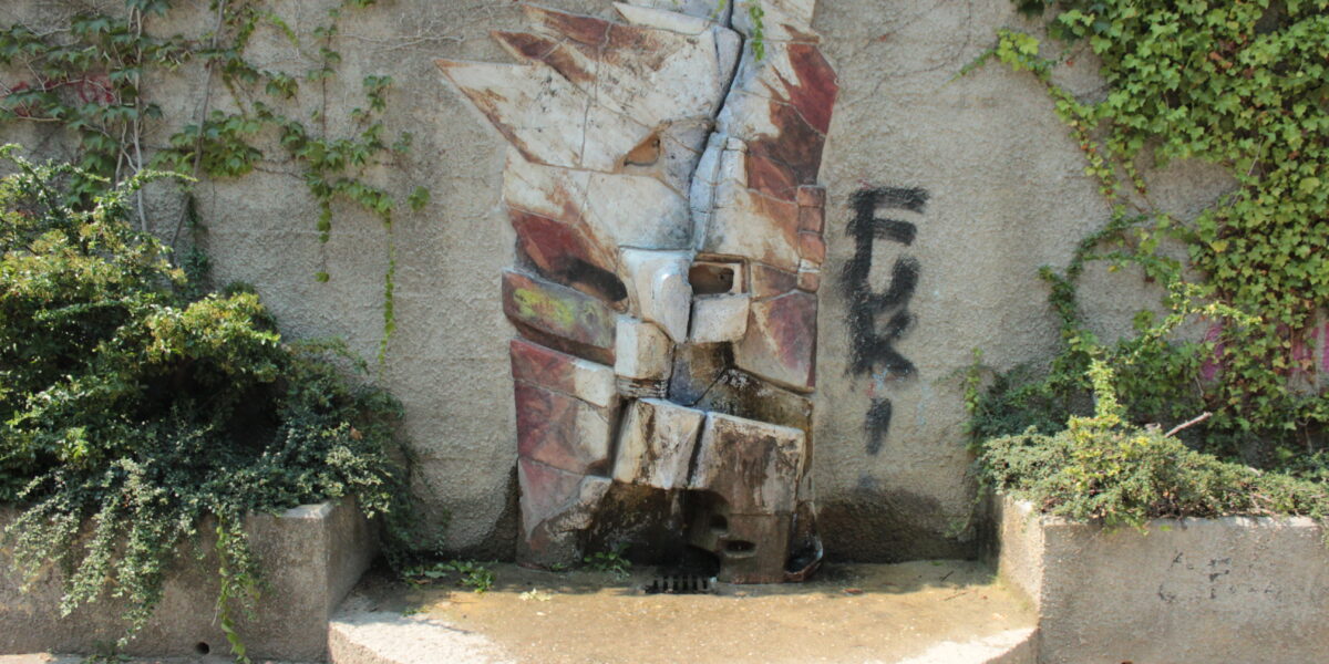 Ein Steinbrunnen mit geometrischem Muster ist in eine Betonmauer eingelassen, die von Grünpflanzen und Ranken flankiert wird. An der Wand rechts neben dem Brunnen befindet sich ein schwarzes Graffiti mit der Aufschrift "FUK!".