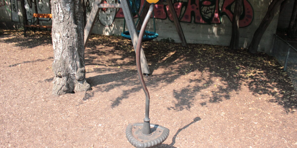 Eine Metallskulptur mit einem gewundenen Stiel und einer gelb-orangefarbenen blütenähnlichen Spitze steht auf einem schmutzigen Boden in der Nähe von Bäumen und einer mit Graffiti beschmierten Wand.
