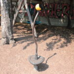 Eine Metallskulptur mit einem gewundenen Stiel und einer gelb-orangefarbenen blütenähnlichen Spitze steht auf einem schmutzigen Boden in der Nähe von Bäumen und einer mit Graffiti beschmierten Wand.