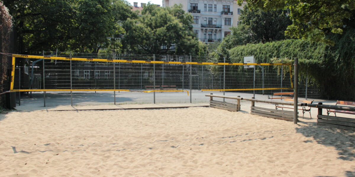 Ein sandiges Volleyballfeld wird von Holzbänken und einem Metallzaun eingefasst, im Hintergrund stehen Bäume und ein Wohnhaus.