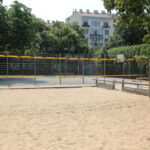 Ein sandiges Volleyballfeld wird von Holzbänken und einem Metallzaun eingefasst, im Hintergrund stehen Bäume und ein Wohnhaus.