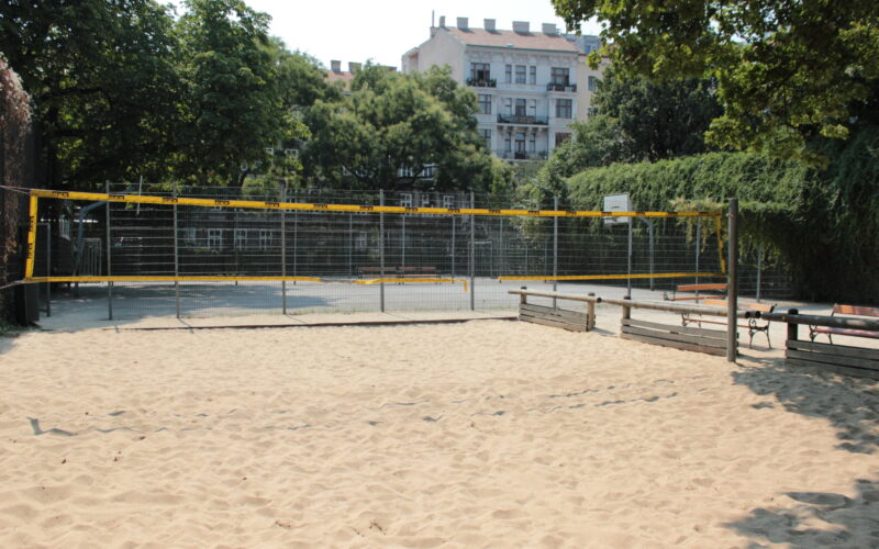Ein sandiges Volleyballfeld wird von Holzbänken und einem Metallzaun eingefasst, im Hintergrund stehen Bäume und ein Wohnhaus.