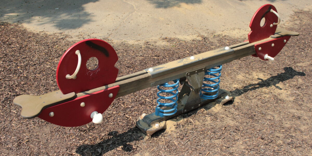Eine rote und braune gefederte Wippe mit tierförmigen Enden auf einem Spielplatz mit Holzspänen und Beton.