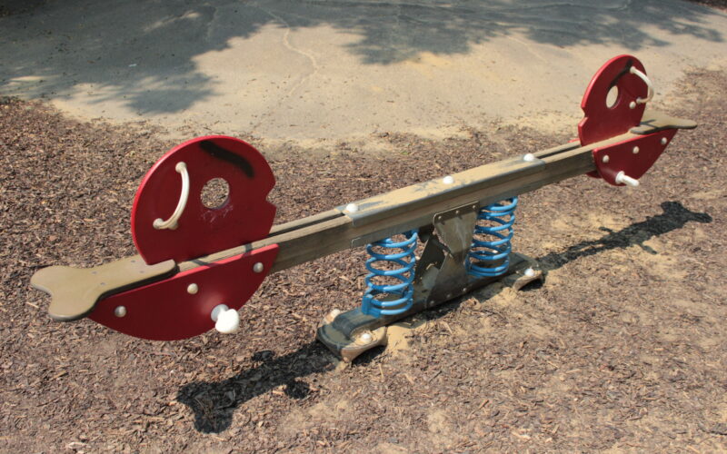 Eine rote und braune gefederte Wippe mit tierförmigen Enden auf einem Spielplatz mit Holzspänen und Beton.