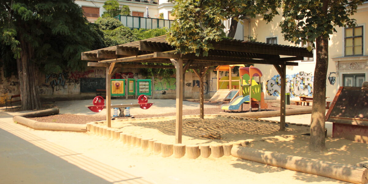Ein Spielplatz mit einer hölzernen Schattenstruktur, Sandkasten, Rutschen, Wippe und Bänken, umgeben von Bäumen und Gebäuden mit Graffiti an den Wänden.