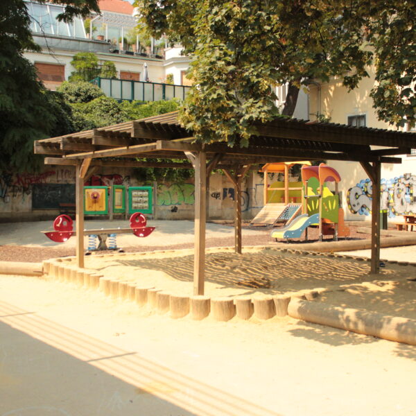 Ein Spielplatz mit einer hölzernen Schattenstruktur, Sandkasten, Rutschen, Wippe und Bänken, umgeben von Bäumen und Gebäuden mit Graffiti an den Wänden.