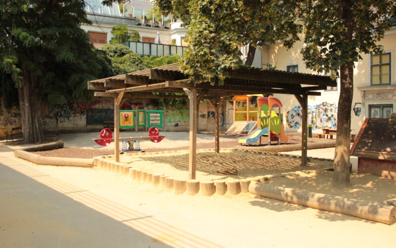 Ein Spielplatz mit einer hölzernen Schattenstruktur, Sandkasten, Rutschen, Wippe und Bänken, umgeben von Bäumen und Gebäuden mit Graffiti an den Wänden.