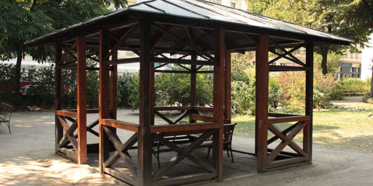 Ein hölzerner Pavillon mit einem Metalldach steht in einem Park, umgeben von Bäumen, Bänken und nahe gelegenen Gebäuden im Hintergrund.
