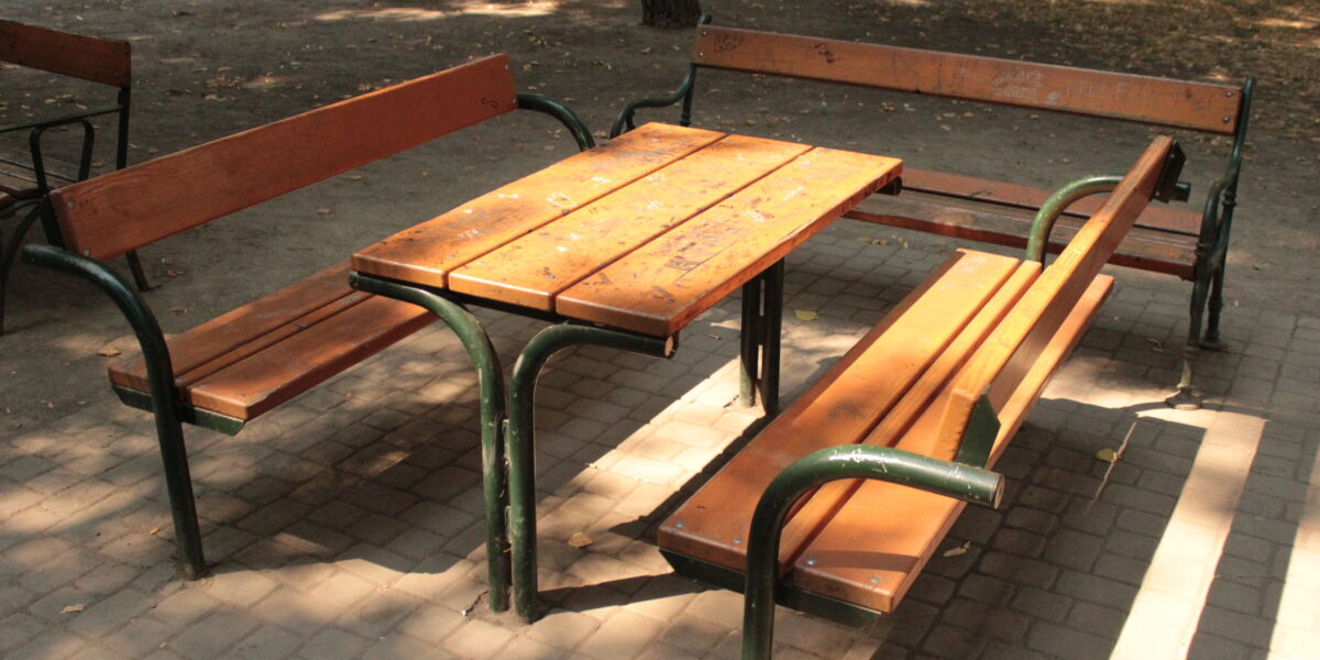 Ein hölzerner Picknicktisch mit an drei Seiten angebrachten Bänken, der auf einer gepflasterten Fläche in einem Park mit Bäumen und trockenem Laub in der Nähe steht.
