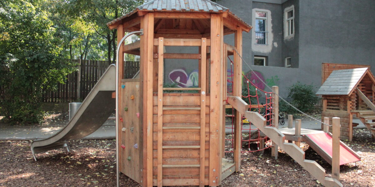 Ein Spielplatz aus Holz mit einer Rutsche, einer Kletterwand, einer Seilbrücke und einer Treppe, der im Freien auf Mulch liegt, mit Bäumen und Gebäuden im Hintergrund.